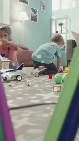 sister and brother decorate roof of wooden house with colorful blocks. children sit on floor in nursery view through clearance in bunch of pencils in glass