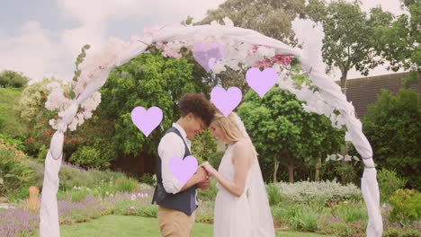 animation of lilac hearts and happy diverse couple holding hands under wedding arch in garden