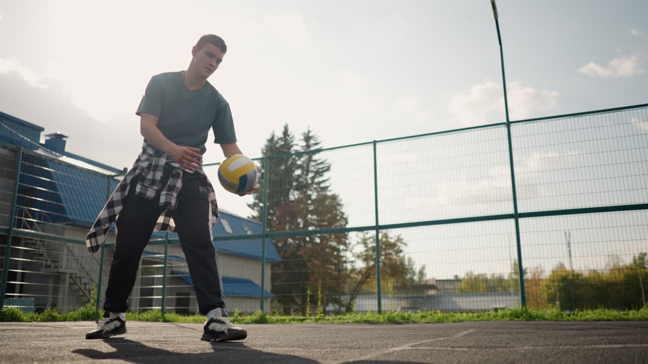 hombre en la cancha de voleibol dejando caer la pelota en el suelo, viendo la pelota rebotar, la cancha presenta cercas y vegetación en el fondo con el edificio en la distancia
