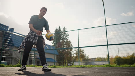 man in volleyball court dropping volleyball on ground, watching ball bounce, court features fencing and greenery in background with building in the distance