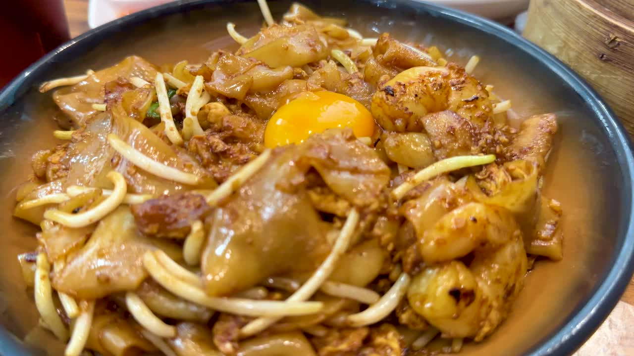 A close-up camera pan reveals a bowl of fried noodles with egg yolk, surrounded by dim sum baskets and Cantonese dishes under bright indoor lighting