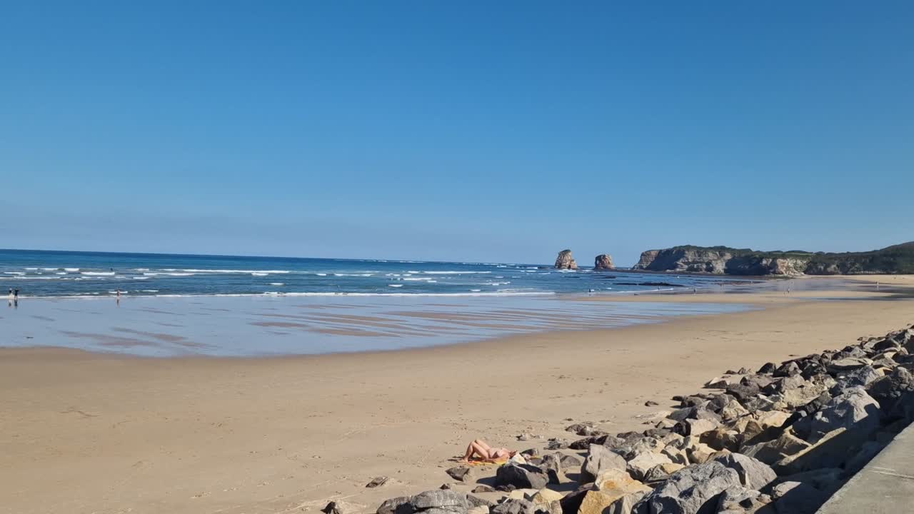 A bright autumn midday scene at Hendaye Beach, on the French Basque coast, showing gentle waves, surfers enjoying the ocean, and a calm blue sky