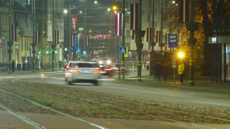 Timelapse-of-city-rush-hour-traffic-on-the-street-of-Liepaja,-traffic-light-streaks,-tram-rails-with-fast-moving-trams,-pedestrians-waiting-for-the-public-transport,-Latvian-flag,-distant-medium-shot