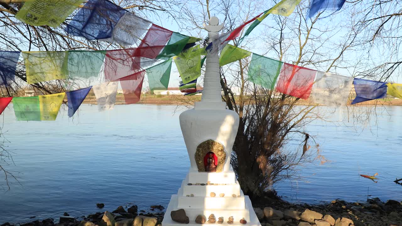 Closeup of small Buddhist stupa ornament on the edge of the river IJssel in Zutphen surrounded by trees and vegetation with colorful flags waving gentle in the wind and water wrinkling in the back