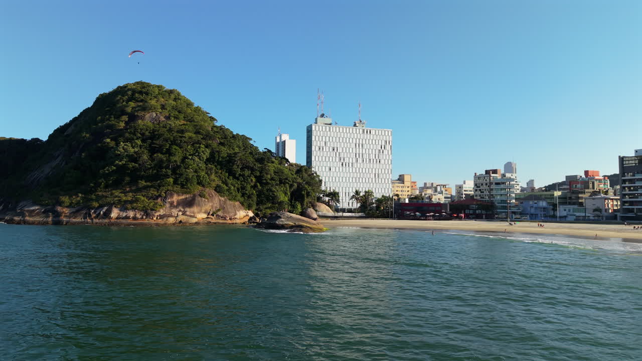 A beautiful aerial drone shot reveals the city beach and skyline of Caiobá, Brazil, with a green mountain and a person paragliding in the distance on a sunny day