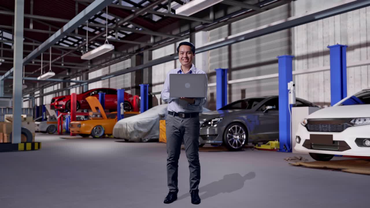 Corpo inteiro de um profissional asiático em pé com seu laptop em uma oficina de conserto de automóveis. Ele está olhando para a câmera com um sorriso.
