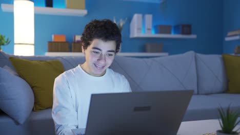 un joven adolescente feliz trabajando en una computadora portátil por la noche en casa.