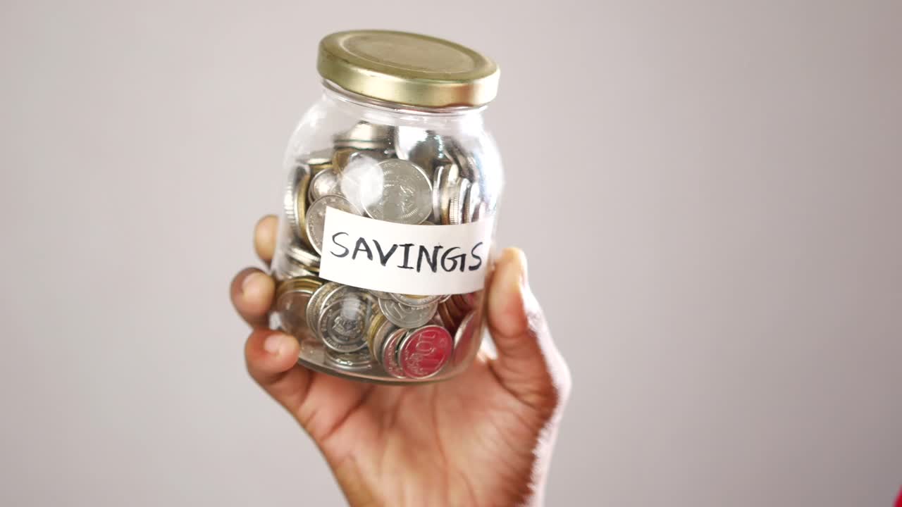 Hand holding a glass jar filled with coins labeled savings