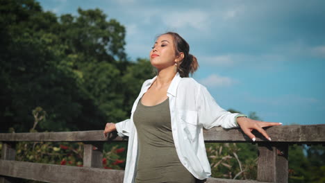 Frau-Genießt-Sonnigen-Tag,-Lehnt-Sich-Gegen-Ein-Holzgeländer-Mit-Einer-üppigen-Landschaft-Und-Blauem-Himmel-Im-Hintergrund,-Selbstbewusstes-Auftreten-Und-Entspannte-Haltung-Spiegeln-Harmonische-Mischung-Aus-Natur-Und-Stil-Wider