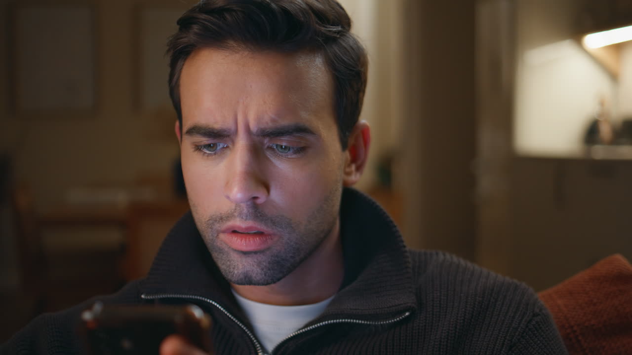 Frowned guy browsing mobile phone app sitting living room couch closeup