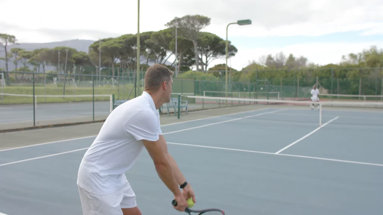 Caucasian male tennis player serving ball to opponent on outdoor tennis court in slow motion