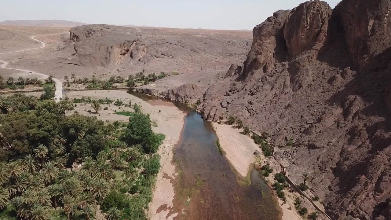 Riprese aeree con drone di un fiume che scorre tra scogliere aspre e una fitta oasi di palme con strutture di villaggi vicini in Marocco. Forte contrasto tra elementi naturali e umani, ideale per viaggi, documentari.