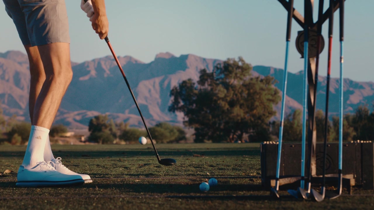 Un primo piano ravvicinato di un golfista che colpisce la palla al campo pratica in slow motion con uno sfondo di montagne desertiche.