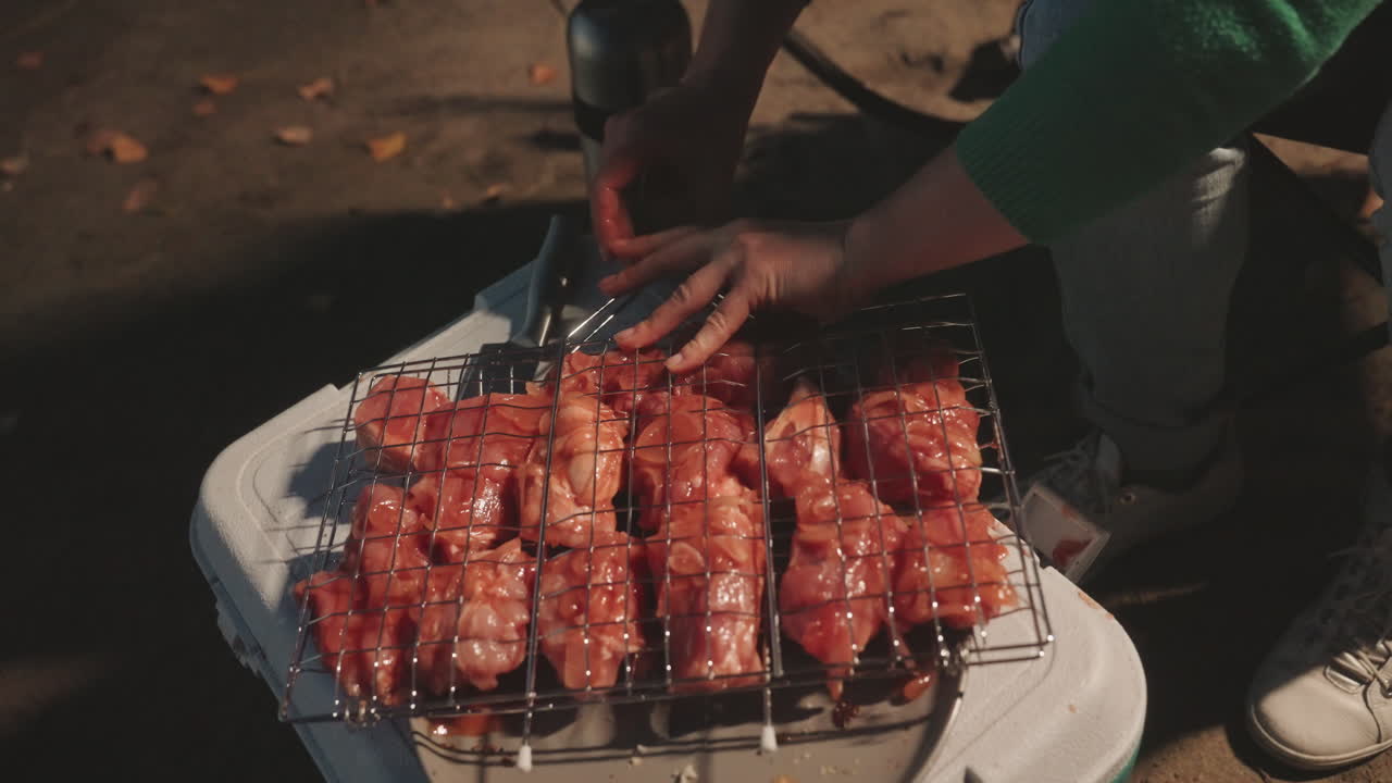 Primo piano di mani da campeggio che posizionano spiedini crudi, condendo e sistemando la carne su una griglia a rete sopra una griglia portatile posta su un frigo portatile, dita che premono gli spiedini nell'apposito supporto, cospargendo sale e pepe, regolando la spaziatura.