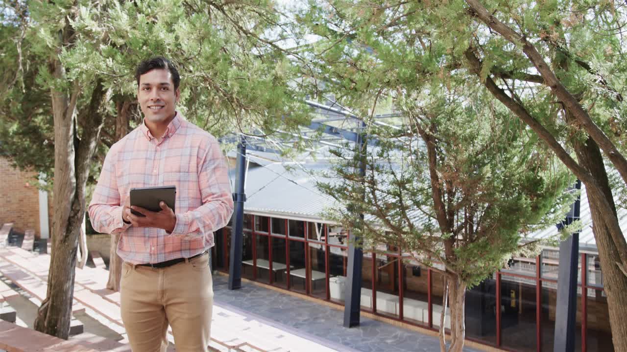 Feliz professor birracial segurando um tablet digital em pé perto de uma escola primária, espaço para texto