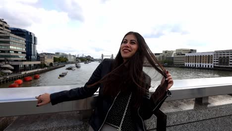 pretty young woman sitting on the london bridge and touching her hair with a smile on her face. slow motion tower bridge at the distance