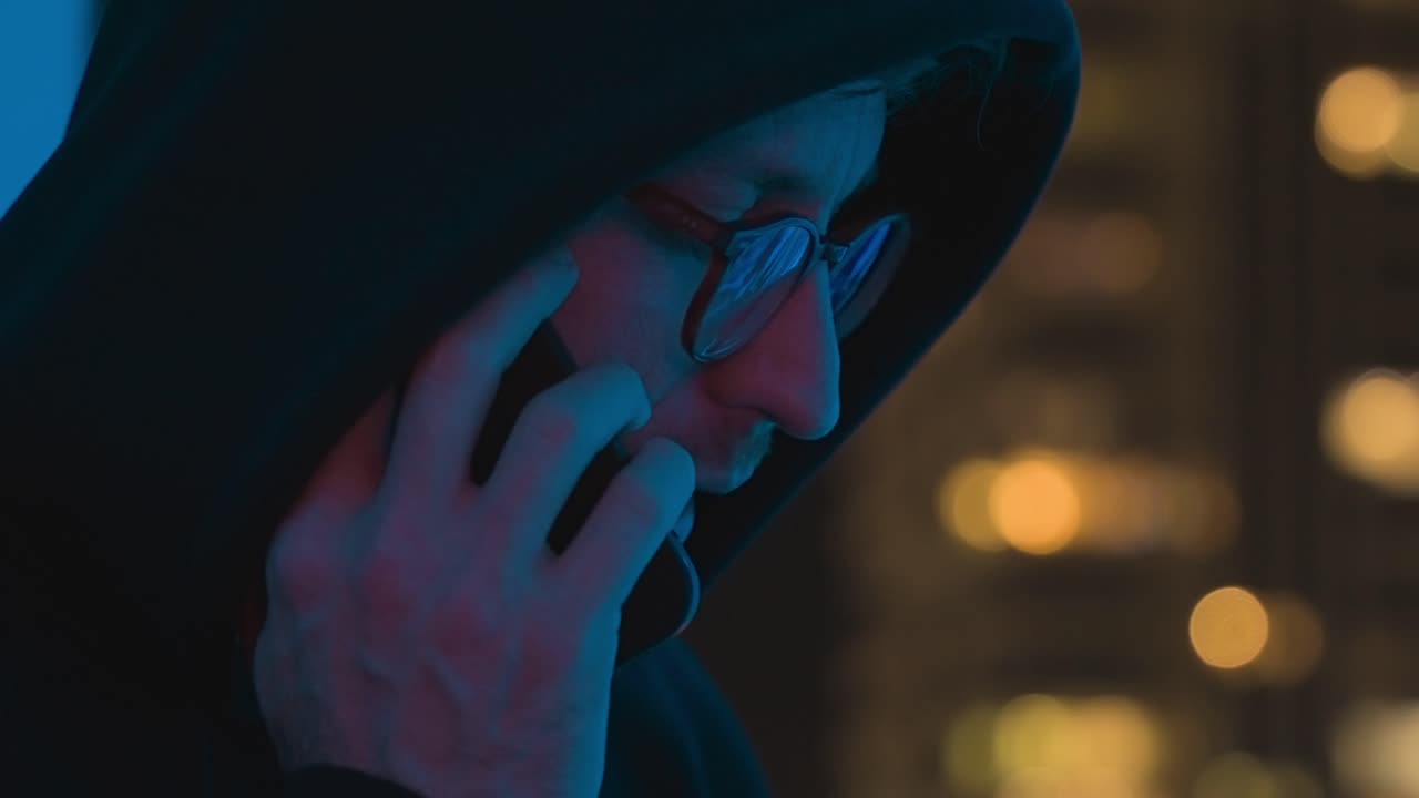 Uomo con felpa nera con cappuccio o hacker che parla al telefono cellulare di notte vicino a una finestra con vista su edifici cittadini. Cospirazione di corruzione o frode. Attività illegale o operazione di insabbiamento criminale. Spia del dark web.