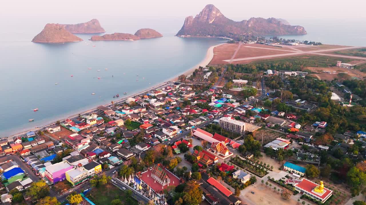 Aerial descend over Prachuap Khiri Khan Thailand coastal city, oceanfront buildings, mountains, and stunning blue waters contrasting against vibrant gold red temples