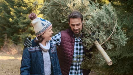 smiling dad and teenage son carrying big christmas tree