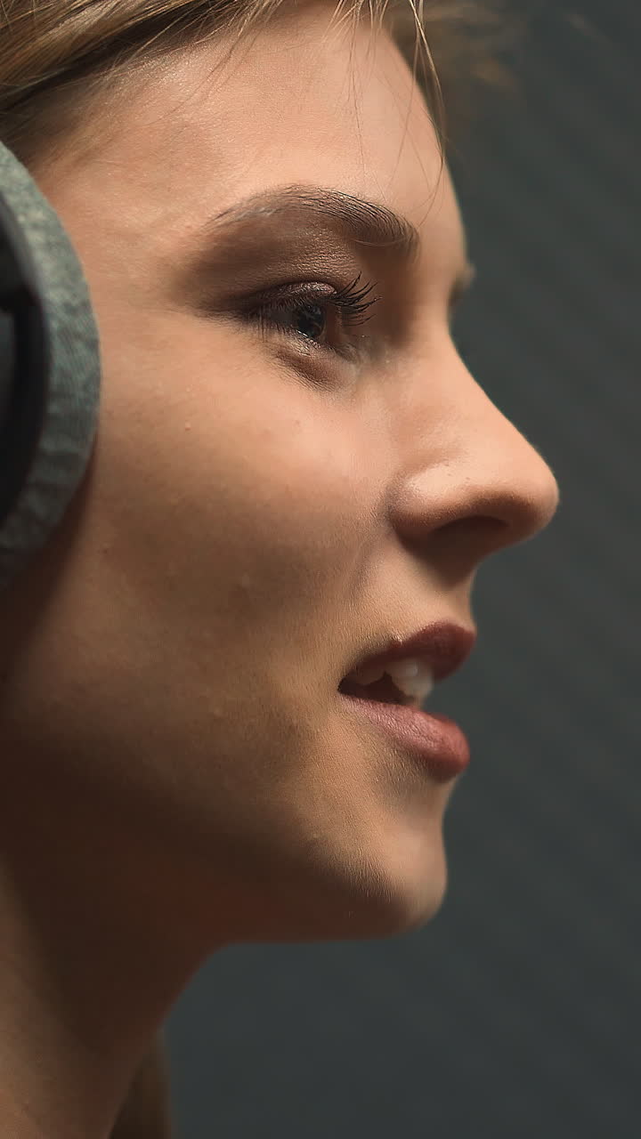 Pretty happy young woman with large black headphones sings popular song with contemporary microphone in recording studio close side view