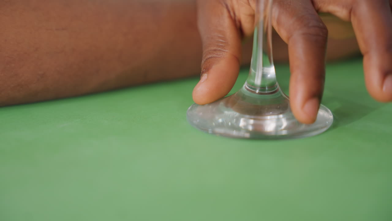 Black hands hold wine glass, closeup on fingertips resting on glass base on green table, intimate date moment with nervous touch, evening bistro lighting, subtle reflection on stem, gentle
