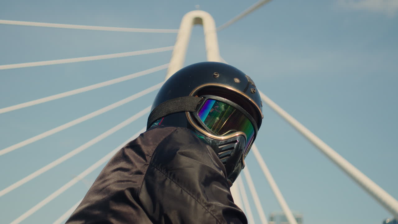 Person in a motorcycle helmet with iridescent goggles looking at a modern bridge