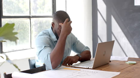 frustrated businessman working on laptop and reviewing blueprints in office