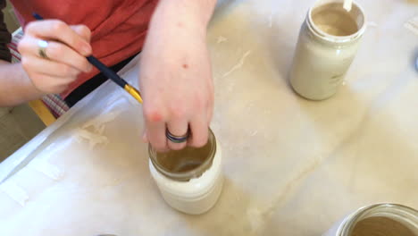 teenage boy sitting on table painting mason jars with white paint, top view and camera going down closer to action