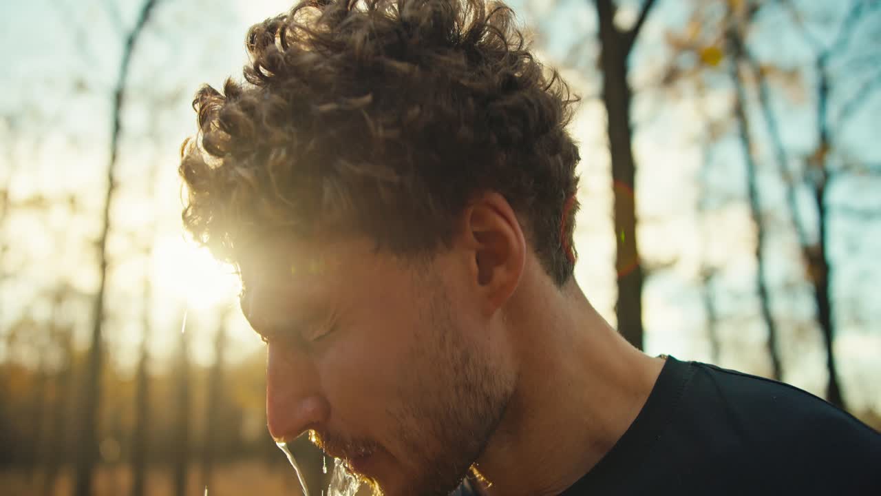 primer plano un hombre cansado con el cabello rizado y una barba vierte agua en su cara de una botella de deportes gris para refrescarse después de una carrera dura en el bosque de otoño. un hombre con un uniforme deportivo negro toma un tiempo antes de correr y se refresca en un bosque de otoño soleado