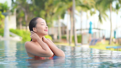 Young-brunette-woman-standing-in-the-swimming-pool-wearing-bikini,-touching-her-wet-hair-rising-her-face-up-with-delight-and-opening-her-eyes