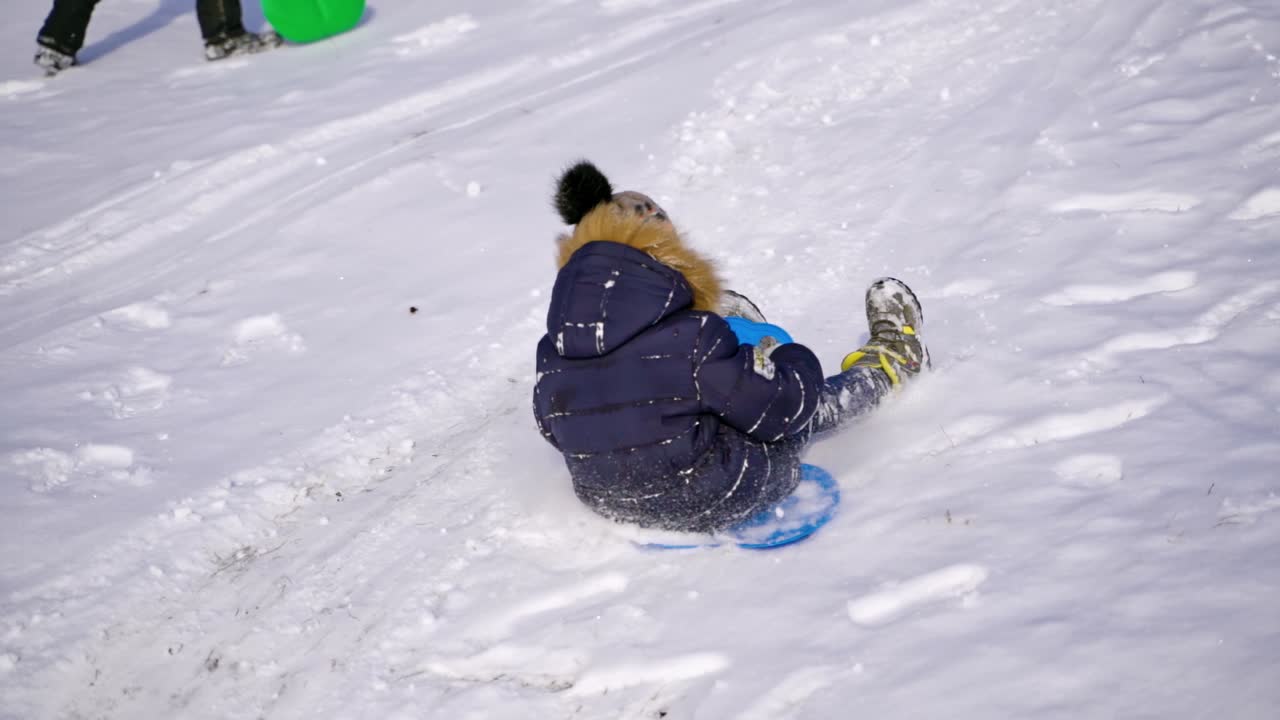 Boy on a snow slides. Little child sledding on plastic sled happily from the white hill in winter. Slow motion.