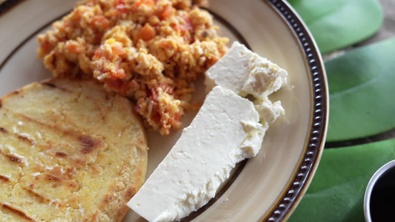 Cận cảnh đĩa bữa sáng Colombia đầy màu sắc gồm trứng bác với cà chua, bánh arepa và phô mai trắng tươi dưới ánh sáng ban mai dịu nhẹ. Sự kết hợp hoàn hảo giữa hương vị và kết cấu