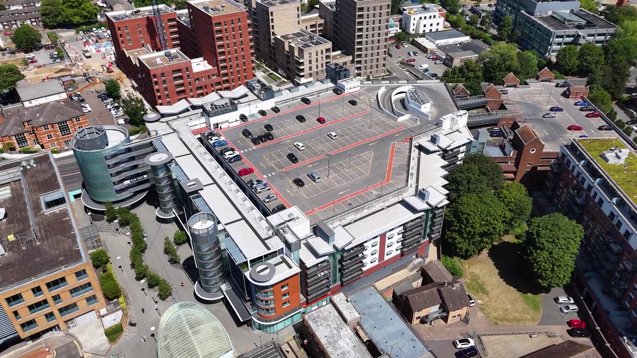 Rooftop car park with red marked bays crowns an office building with circular glass stair towers, forming a strong geometric pattern from above