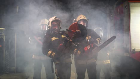 group of firefighters wearing full equipment, oxygen masks, and emergency rescue tools, circular hydraulic and gas saw, axe, and sledge hammer. smoke and fire trucks in the background.