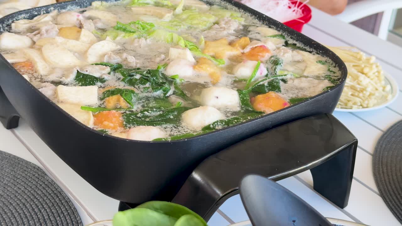 A vibrant hotpot simmers with vegetables, meat, and fishballs in an outdoor setting, highlighting communal dining and home cooking
