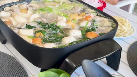 a vibrant hotpot simmers with vegetables, meat, and fishballs in an outdoor setting, highlighting communal dining and home cooking