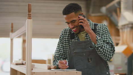 male apprentice working as carpenter in furniture workshop talking on mobile phone