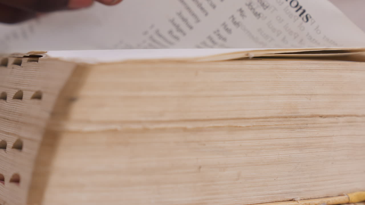 Pagine di un libro spesso sfogliate a mano, primo piano dei bordi delle pagine e della rilegatura consumati, rapido sfogliare con il pollice e gesto deliberato, contesto di un tavolo da biblioteca, trama archivistica e intensità della ricerca, sottile luce ambientale.