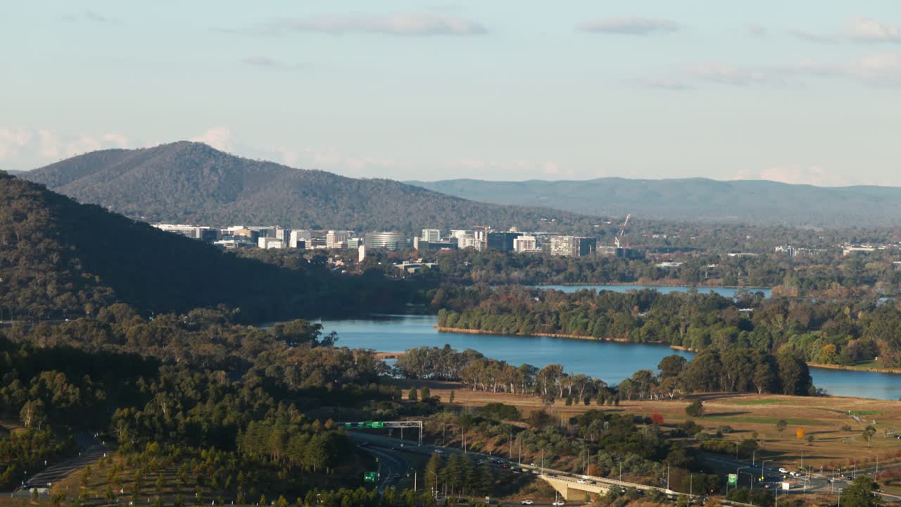 A high vantage point captures Canberra CBD and surrounding ranges, with autumn foliage softening the urban features and roadways.