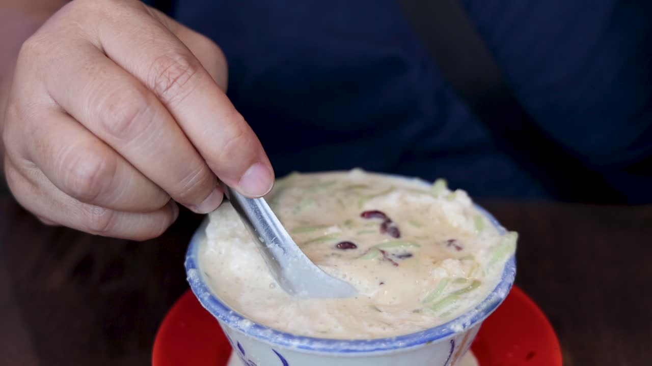A person uses a spoon to mix and scoop cendol dessert in a ceramic bowl, under soft indoor lighting, with a close-up, steady camera angle