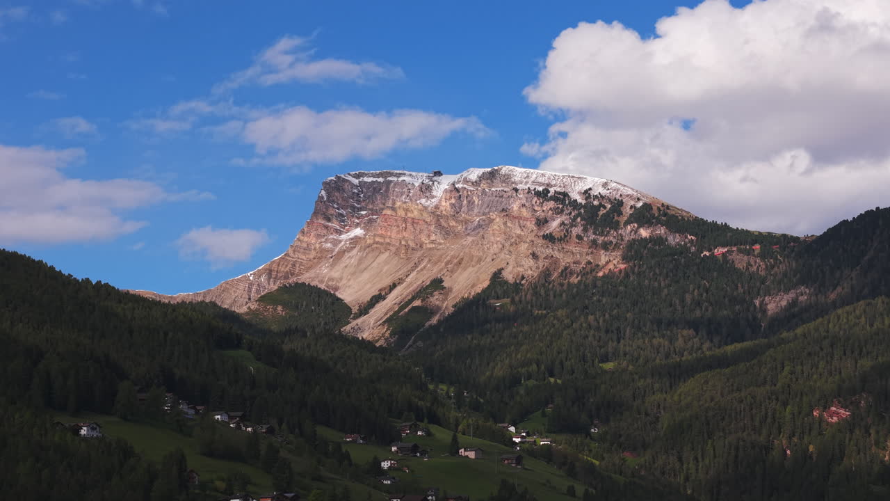 Drone footage of a rocky mountain peak with traces of snow on top, surrounded by green forested slopes and scattered houses in the Dolomites, Italy