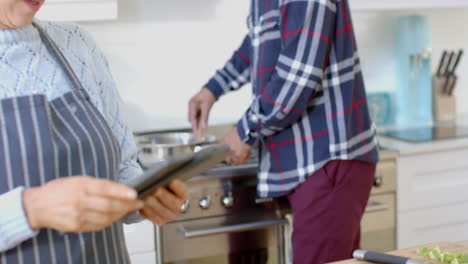 Feliz-Pareja-Birracial-Mayor-Cocinando-Y-Usando-Tableta-En-La-Cocina,-Cámara-Lenta