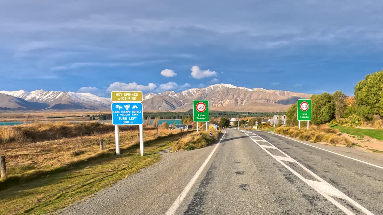 A vehicle travels along a rural highway toward Lake Tekapo, passing road signs, autumn foliage, and snow-capped mountains under bright daylight with steady camera movement