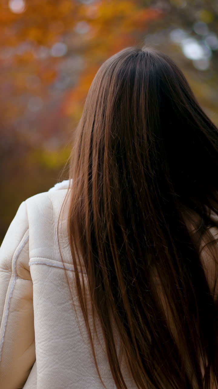 Long-haired brunette woman walking in the park. Lady turns around looks into camera holding a phone in her hand. Autumn nature backdrop. Vertical video