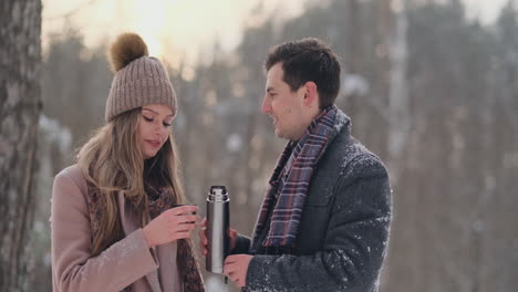 pareja feliz sosteniendo tazas de té calientes sobre el paisaje invernal. una joven pareja enamorada en unas vacaciones de invierno, parada junto a un árbol y bebiendo una taza de té caliente