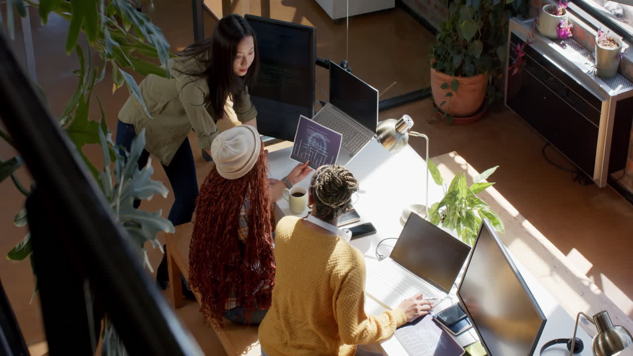 Diverse female team collaborating on coding project in modern office with laptops and documents