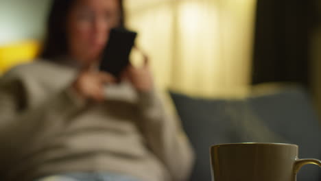 woman spending evening at home sitting on sofa with mobile phone scrolling through internet or social media with hot drink in foreground