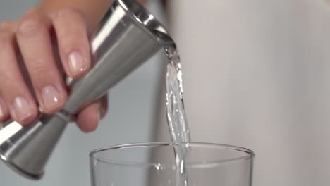 Woman-bartender-pouring-transparent-alcoholic-liquid-from-metal-jigger-into-clear-class-close-up-slow-motion