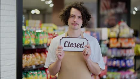 Portrait-of-a-sad-brunette-guy-in-an-apron-who-holds-in-his-hands-a-sign-with-the-inscription-closed-in-a-supermarket