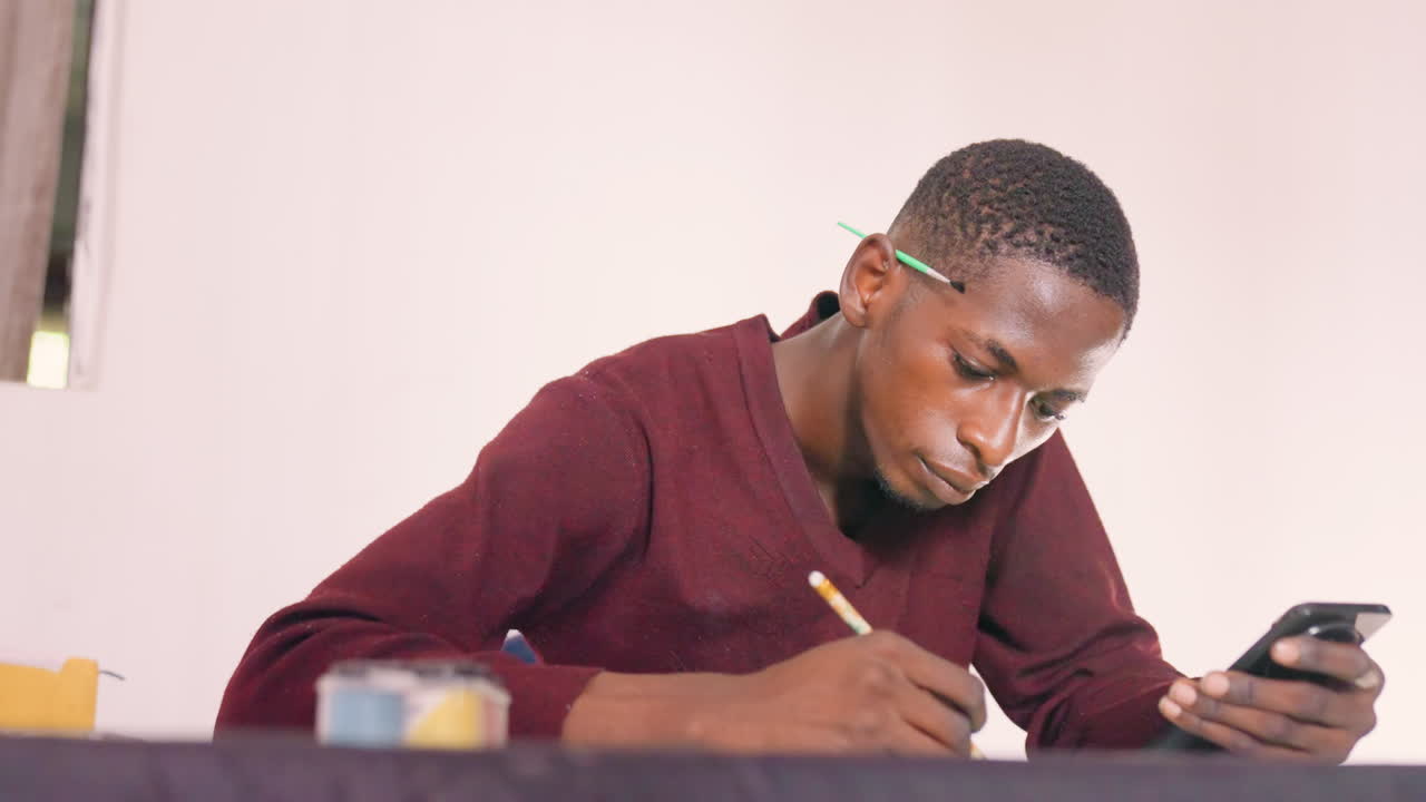 Artista Masculino Negro Pintando com Referência no Telefone, Equilibrando Esboço Rápido e Notas de Cores na Mesa, Postura Concentrada em Suéter Bordô, Cena Interna de Mesa com Potes de Tinta, Humor Determinado e Produtivo